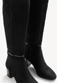 Bottes noires au-dessus du genou en daim avec une texture lisse, dotées d'une fermeture éclair et d'un accent noir brillant autour de la cheville.