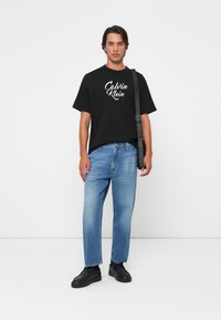 Hombre vistiendo una camiseta negra de Calvin Klein, jeans azul claro, zapatos negros y llevando una bolsa de hombro negra contra un fondo blanco.