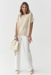 Maglione beige a maglia con maniche corte, pantaloni bianchi a palazzo e una borsa beige chiaro. Scarpe con tacco alto bianche con cinghie alla caviglia.