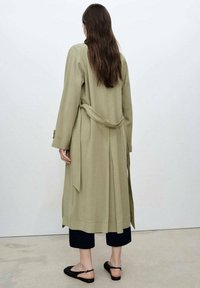 Długi płaszcz oliwkowozielony z wiązaniem w talii, długimi rękawami i prostym krojem; zestawiony z ciemnymi, krótko przyciętymi spodniami i czarnymi płaskimi butami.