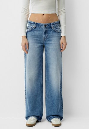 Woman wearing wide-leg high-waist blue jeans, white long-sleeve crop top, and white sneakers, standing against a plain light background.