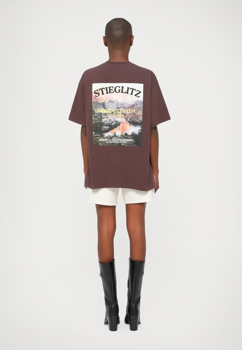 Camiseta oversized marrón con un gráfico de paisaje montañoso; presenta texto en blanco y amarillo. Combinada con shorts blancos y botas altas negras.