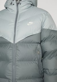 Casaco puffer cinza com fecho éclair na frente, capuz e design Chevron. Apresenta um logo da Nike no peito e um bolso lateral com fecho.
