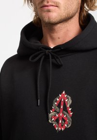 Sort hættetrøje med et tekstureret rødt og guld design med spidse detaljer på forsiden. Snore i halsudskæringen giver mulighed for justerbar pasform.
