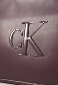 Sac en cuir violet avec un logo "ck" en relief dans une finition brillante. Texture lisse avec un grain subtil et des coutures contrastantes.