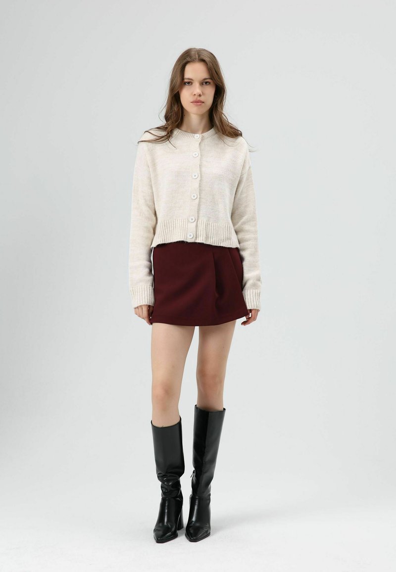 Roomkleurige cropped cardigan met knopen, gecombineerd met een bordeauxrode minirok en knielange zwarte leren laarzen met spitse neuzen.