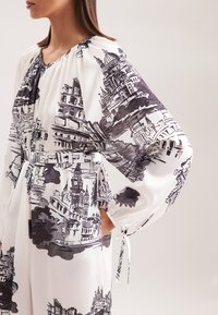 Weißes Kleid mit architektonischem Schwarz-Weiß-Druck, lange Ärmel, geraffter Ausschnitt und Taillenzugdetails. Weicher, fließender Stoff.