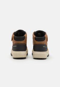 Bottines en cuir brun avec accents noirs, surface texturée, sangle ajustable et semelle beige. Présente un logo sur le talon.