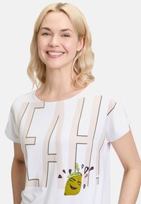 Witte T-shirt met korte mouwen met "YEAH!" in vet lichtbruin lettertype en een cartoon citroenafbeelding met groene bladeren en druppels.