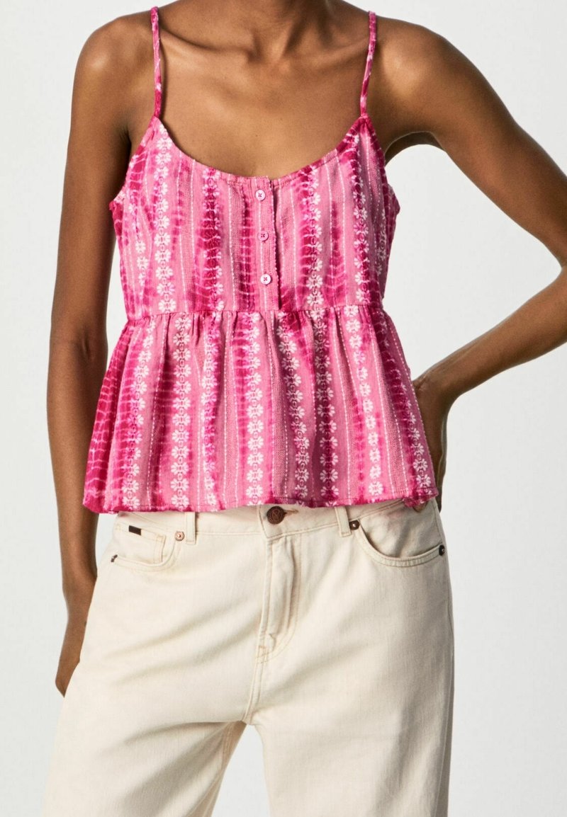 Femme portant un haut rose sans manches à boutons avec des rayures florales blanches et un pantalon beige, debout avec une main sur la hanche.
