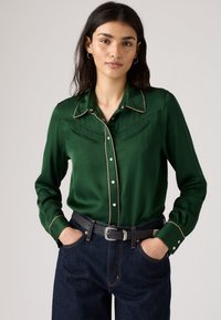 Grünes, seidiges Hemd mit Knopfleiste und weißer Paspelierung, zwei Brusttaschen und einem spitzen Kragen, kombiniert mit dunklen Jeans und einem schwarzen Gürtel.