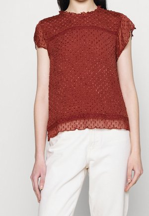 Frau trägt eine durchsichtige, rostrote, kurzärmelige Bluse mit erhobenen Polka-Dots und weißen, hochgeschnittenen Hosen, kurz geschnitten an Hals und Hüften.