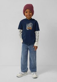 Granatowy t-shirt z napisem "Mniej bałaganu, więcej", nałożony na kremową koszulę z długim rękawem z czarnymi graficznymi akcentami. Jasnoniebieskie dżinsy i białe sneakersy.