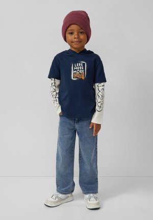 Marineblaues T-Shirt mit dem Text "WENIGER CHAOS MEHR", übereinander getragen mit einem cremefarbenen Langarmshirt mit schwarzen grafischen Akzenten. Hellblaue Jeans und weiße Sneaker.