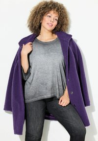 Violetti fleece-takki, jossa on teksturoitu pinta, yhdistettynä harmaaseen ylisuureen paitaan ja tummiin farkkuihin. Takissa on suuria nappia ja kaulus.