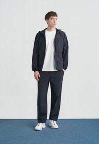 Νέος άνδρας που στέκεται σε μπλε χαλί φορώντας μαύρο σακάκι, λευκό μπλουζάκι, μαύρο παντελόνι και λευκά αθλητικά παπούτσια, κοιτάζει προς το πλάι.