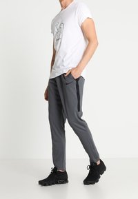 Harmaat urheiluhaalarit, joissa on mustat sivuraitat, yhdistettynä valkoiseen t-paitaan, jossa on graafinen painatus. Mustat lenkkarit viimeistelevät asustuksen.