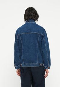 Tumši zilā denim jaka, kurai ir klasiskā apkakle, garas piedurknes un pogu akcenti pie aprocēm un jostasvietas. Vienkāršs aizmugures dizains.