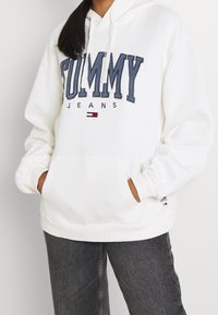 Persona vestida con una sudadera blanca de Tommy Jeans con un logo azul y jeans gris oscuro, con las manos metidas en el bolsillo frontal de la sudadera.