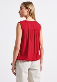 Blouse sans manches rouge avec un détail froncé à l'arrière, fabriquée dans un tissu lisse. Portée avec un pantalon ample de couleur claire.