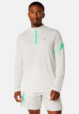 Mann trägt ein weißes, langärmliges Sportoberteil mit grünem Reißverschluss und Logo am Ärmel, dazu passende weiße Shorts mit grünem Logo am linken Bein.