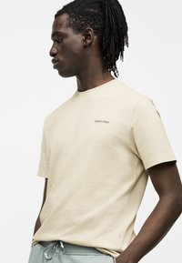 Νέος άνδρας με σκούρα πλεξούδα φοράει μπεζ μπλουζάκι Calvin Klein και ανοιχτό γκρι παντελόνι με κορδόνι, στέκεται με τα χέρια στις τσέπες.