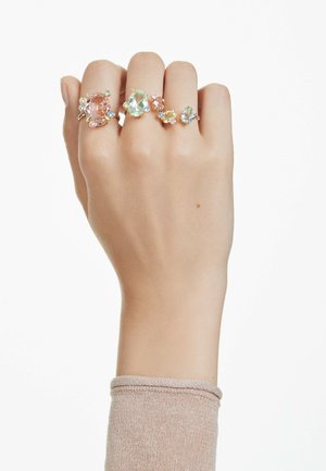 A hand wearing four rings with colorful gemstones, showcasing varying designs and sizes, against a plain background.