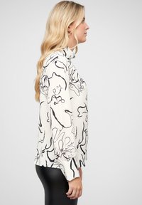 Blouse blanche avec un motif floral abstrait noir, manches longues et col montant ; associée à des leggings noirs. Texture de tissu lisse.