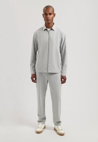 Lys grå jakke med glidelås og matchende bukser i et teksturert stoff, kombinert med hvite og beige joggesko. Enkel, strømlinjeformet design.