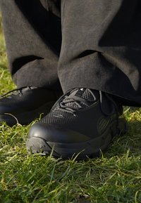 Fekete sportcipők hálós panelek és gumitalpak, lekerekített orrhegyekkel és fűzőkkel, fűre helyezve.