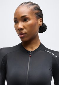 Γυναίκα με πλεξούδες σε χαμηλό κότσο που φοράει μαύρη ποδηλατική φανέλα Castelli με εμπρόσθιο φερμουάρ, κοιτάζει ελαφρώς προς το πλάι.