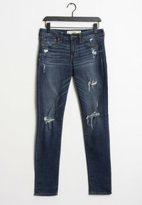 Jeans skinny de mezclilla azul oscuro con múltiples desgastes y bordes deshilachados. Cuenta con cinco bolsillos y cierre de cremallera, colgando de una percha negra.