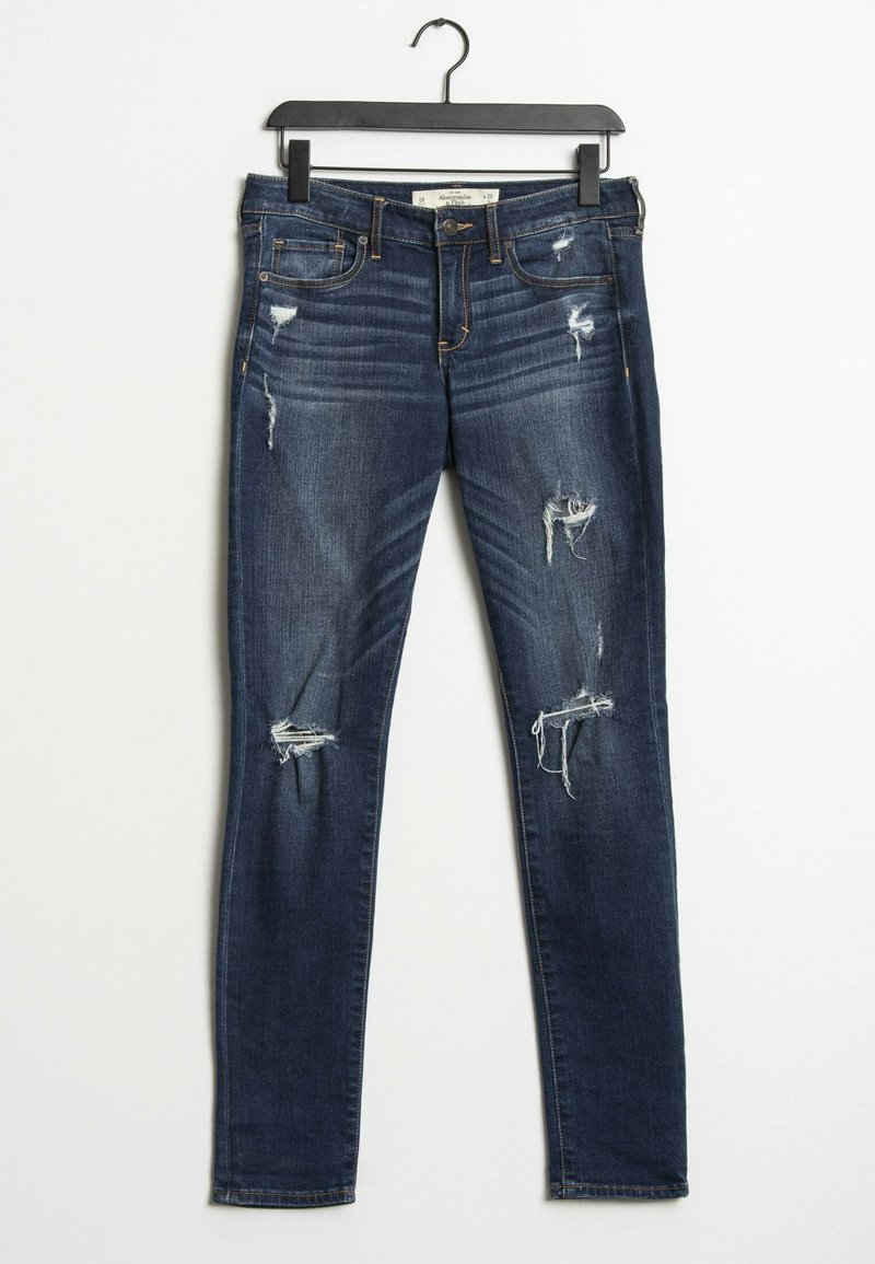 Jeans skinny de mezclilla azul oscuro con múltiples desgastes y bordes deshilachados. Cuenta con cinco bolsillos y cierre de cremallera, colgando de una percha negra.