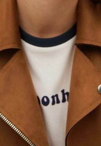 Giacca in suede marrone con zip argentata, indossata sopra una t-shirt bianca con testo blu scuro. Collo arrotondato con rifinitura a contrasto.