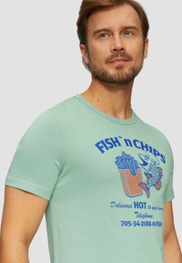 Mintgroene t-shirt met korte mouwen met een graphic van fish and chips in blauw en oranje. Zacht, lichtgewicht materiaal met een ronde hals.