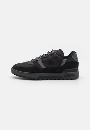 Sort Lacoste sneaker med tekstureret sål, grå øjne og logo på siden og hælen, sat mod en ensfarvet hvid baggrund.
