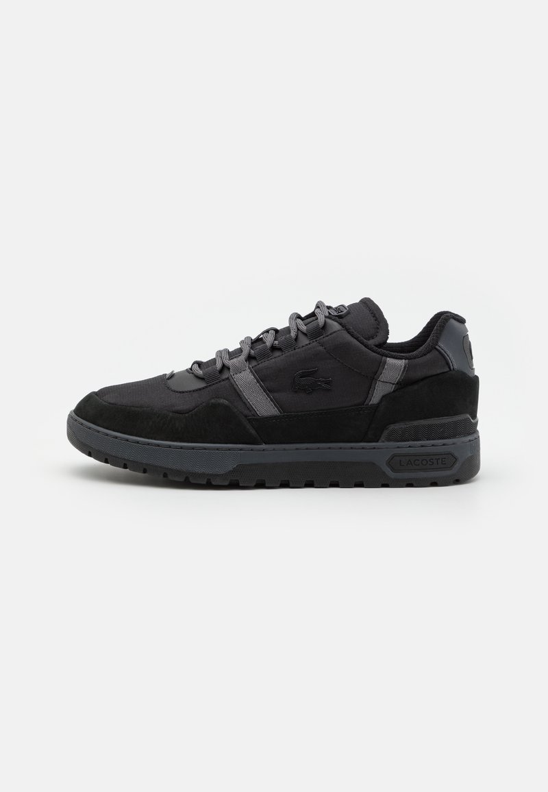 Baskets noires Lacoste avec semelle texturée, œillets gris et logo sur le côté et le talon, sur un fond blanc uni.