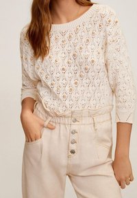 Kremowy sweter dzianinowy z diamentowym wzorem i dekoracyjnymi koralikami, w zestawieniu z beżowymi spodniami o wysokim stanie, z zapięciem na guziki i bocznymi kieszeniami.