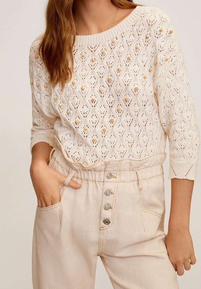 Kremowy sweter dzianinowy z diamentowym wzorem i dekoracyjnymi koralikami, w zestawieniu z beżowymi spodniami o wysokim stanie, z zapięciem na guziki i bocznymi kieszeniami.