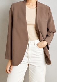 Blazer marrón sin estructura, de tela suave, con un bolsillo en el pecho y solapas anchas, combinado con una camiseta beige y pantalones blancos.