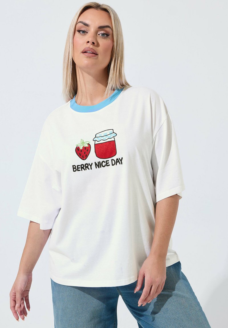 Frau trägt ein weißes, übergroßes T-Shirt mit blauem Kragen und einer Grafik mit Erdbeere und Glas, beschriftet mit "BERRY NICE DAY".