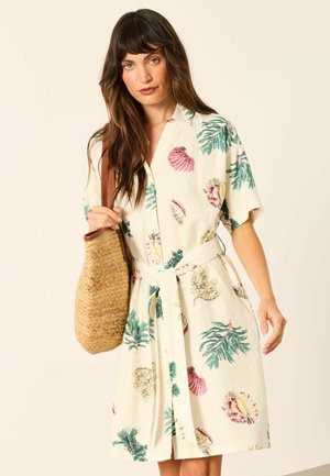 Frau mit langen Haaren trägt ein kurzärmeliges, tailliertes Kleid mit bunten Blatt- und Muschelmotiven und hält eine geflochtene Strohtasche.