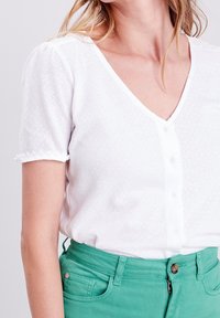 Blusa blanca de manga corta con escote en V y botones en la parte frontal. Tejido texturizado con detalle en los puños de las mangas. Combinada con pantalones verdes de cintura alta.