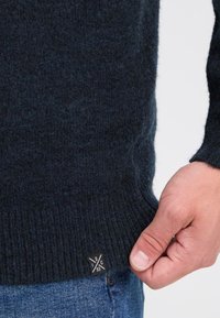 Donkerblauwe trui gemaakt van zacht, textuurvol breiwerk. Heeft een geribde zoom en een klein zwart etiket met witte details. Hand getoond voor schaal.