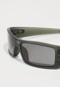 Ochelari de soare verde cu o finisare mată, lentile rectangulare și un cadru robust. Au lentile cu un gradient mai închis și detalii de branding subtile.