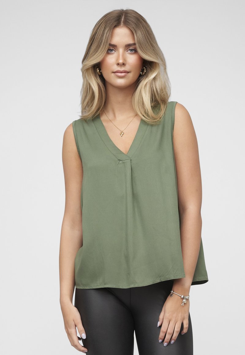 Olivengrønn ermeløs bluse med V-hals, subtil rynkedetalj foran og løs passform, kombinert med svarte leggings.