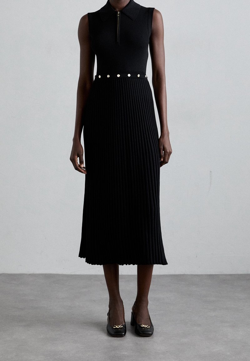 Robe noire sans manches en maille côtelée avec col, fermeture éclair à l'avant et ceinture à boutons argentés, portée avec des talons noirs à bout carré avec un détail chaîne dorée.