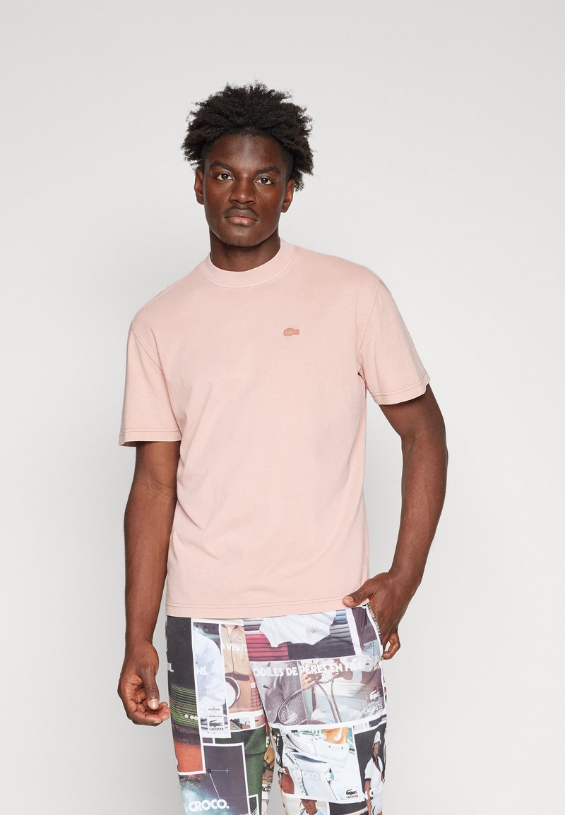 Jeune homme portant une chemise à manches courtes rose clair avec un petit logo et un short multicolore avec des imprimés graphiques, se tenant devant un fond uni.