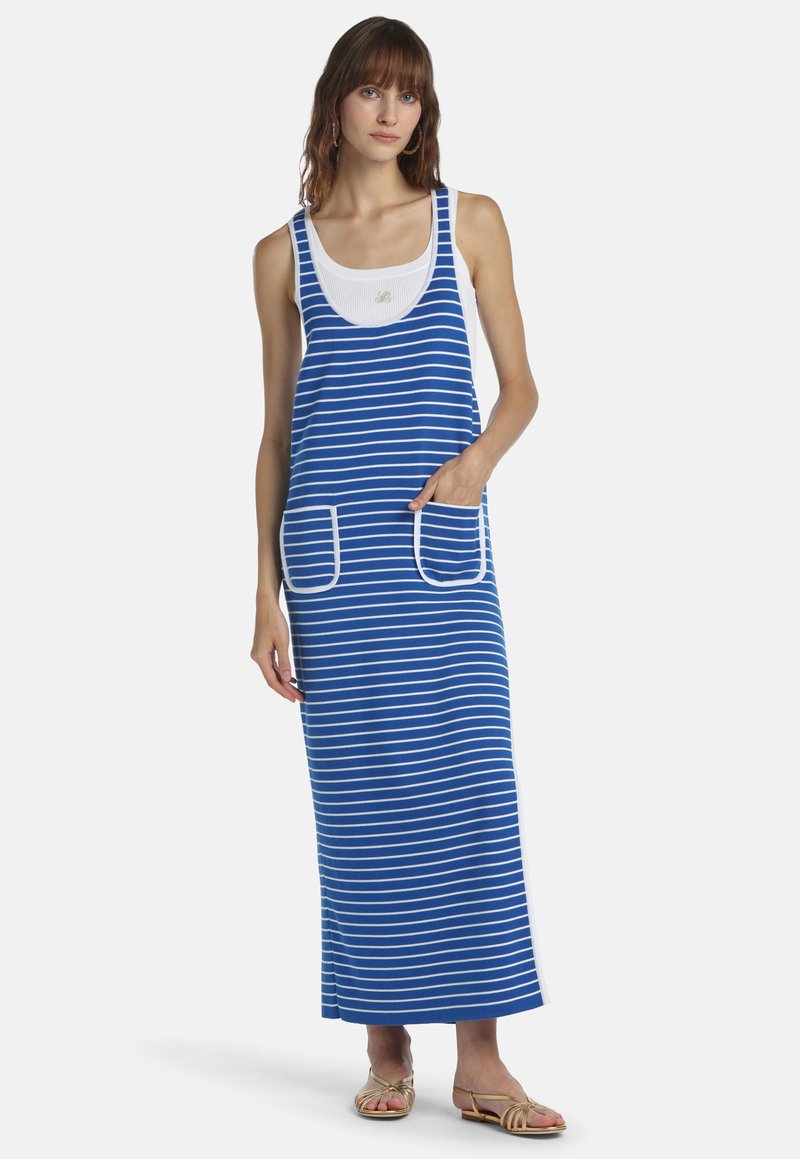 Femme portant une robe maxi sans manches à rayures bleu et blanc avec des poches avant, superposée sur un débardeur blanc côtelé et des sandales beige.