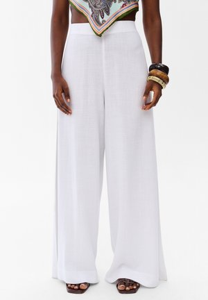 Szerokie białe lniane spodnie noszone z brązowymi sandałami o kwadratowym nosku, kolorową wzorzystą bluzką z chusty oraz nałożonymi bransoletkami na jednym nadgarstku.